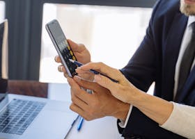Business professionals reviewing financial reports on a smartphone at a desk with papers and a laptop in an office setting.